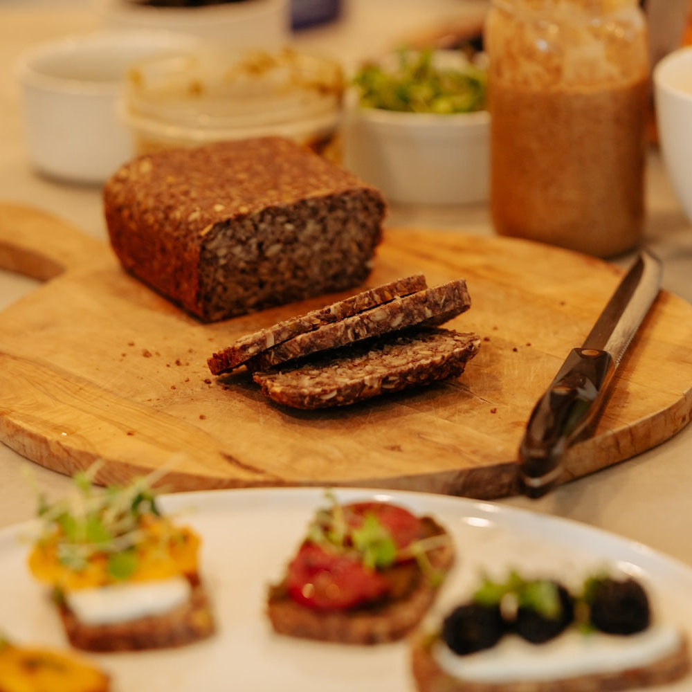 Loaf of bread on a wooden cutting board with sliced bread, a knife, and small appetizers on a plate.